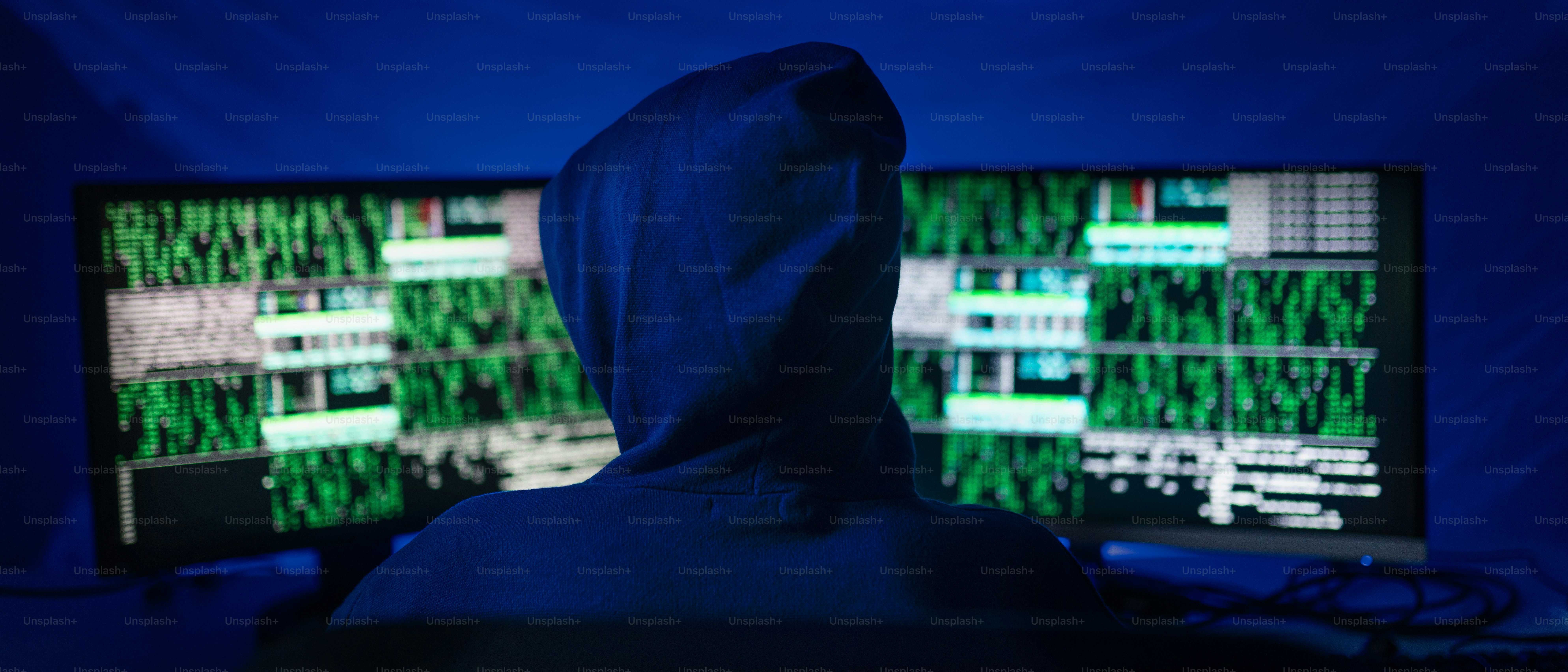 A rear view od hooded hacker by computer in the dark room at night, cyberwar concept